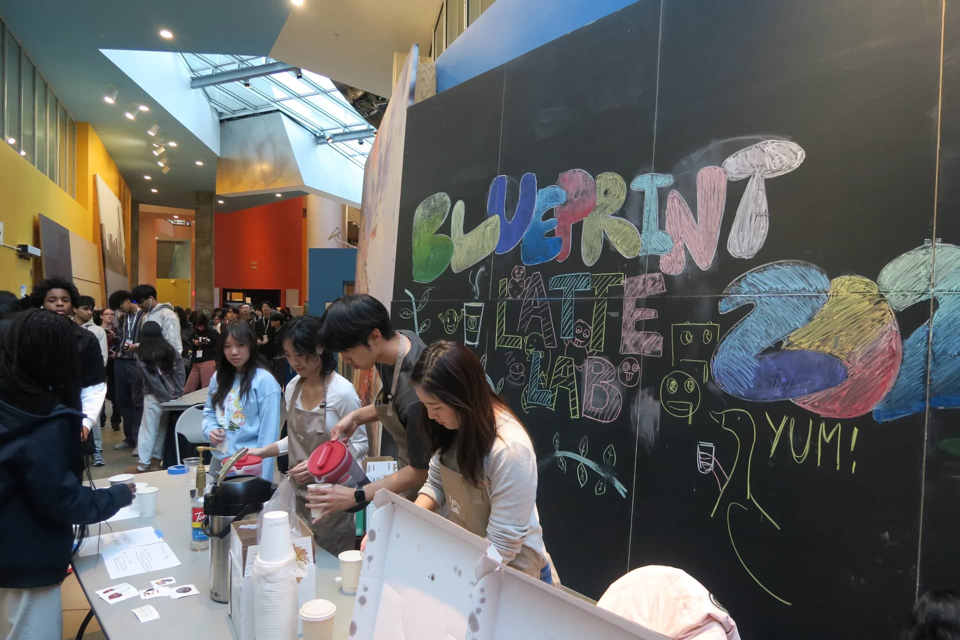Latte Lab volunteers serving drinks beside the colorful Blueprint chalkboard