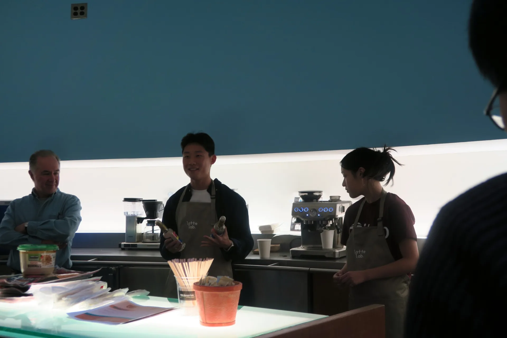 A Latte Lab member in a Latte Lab apron talking with a teammate behind the counter at the CSAIL R&D event, with a coffee maker and espresso machine on the counter
