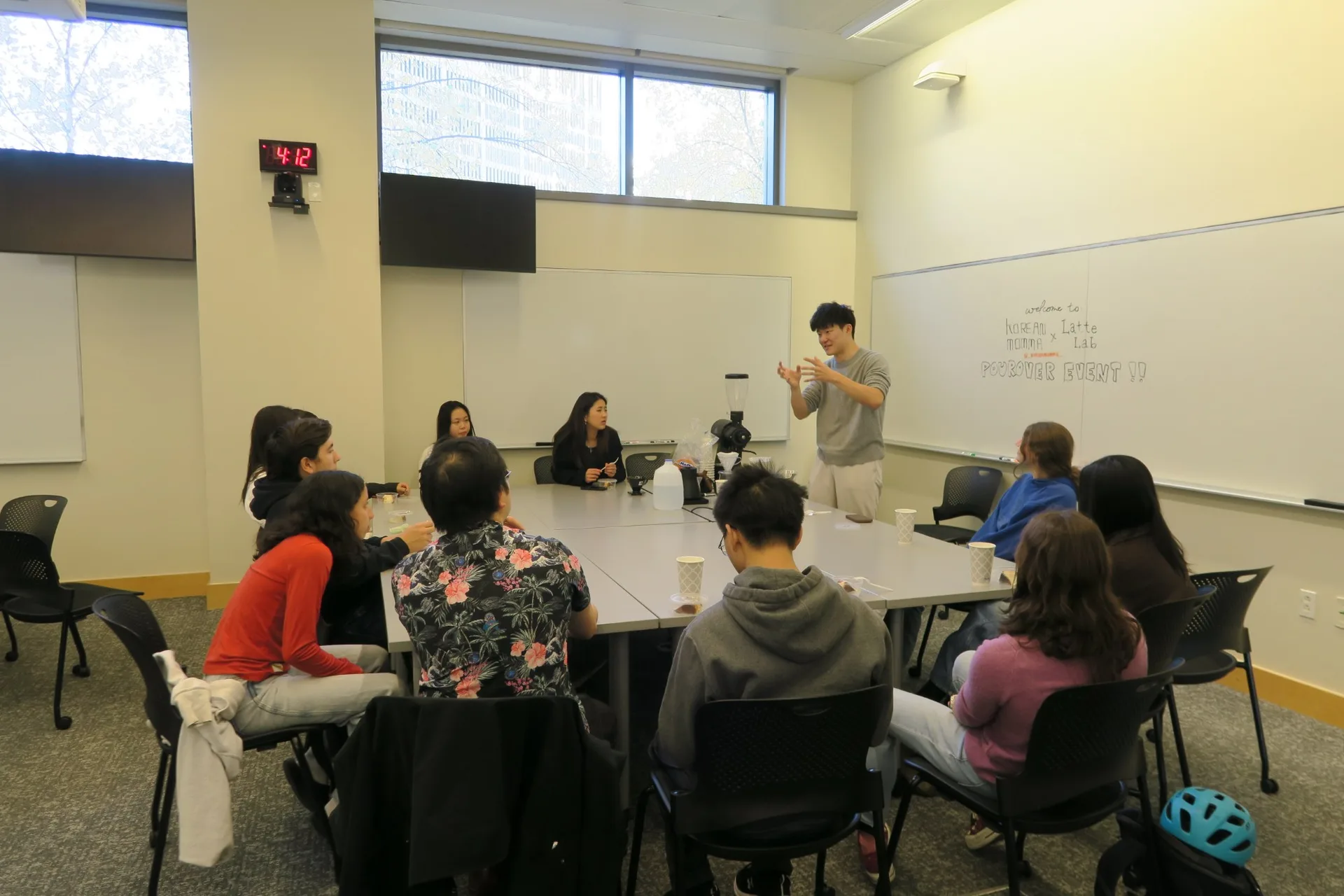 Latte Lab members seated around a table for a Korean Momma x Latte Lab pourover event, with a presenter explaining brewing in front of a whiteboard