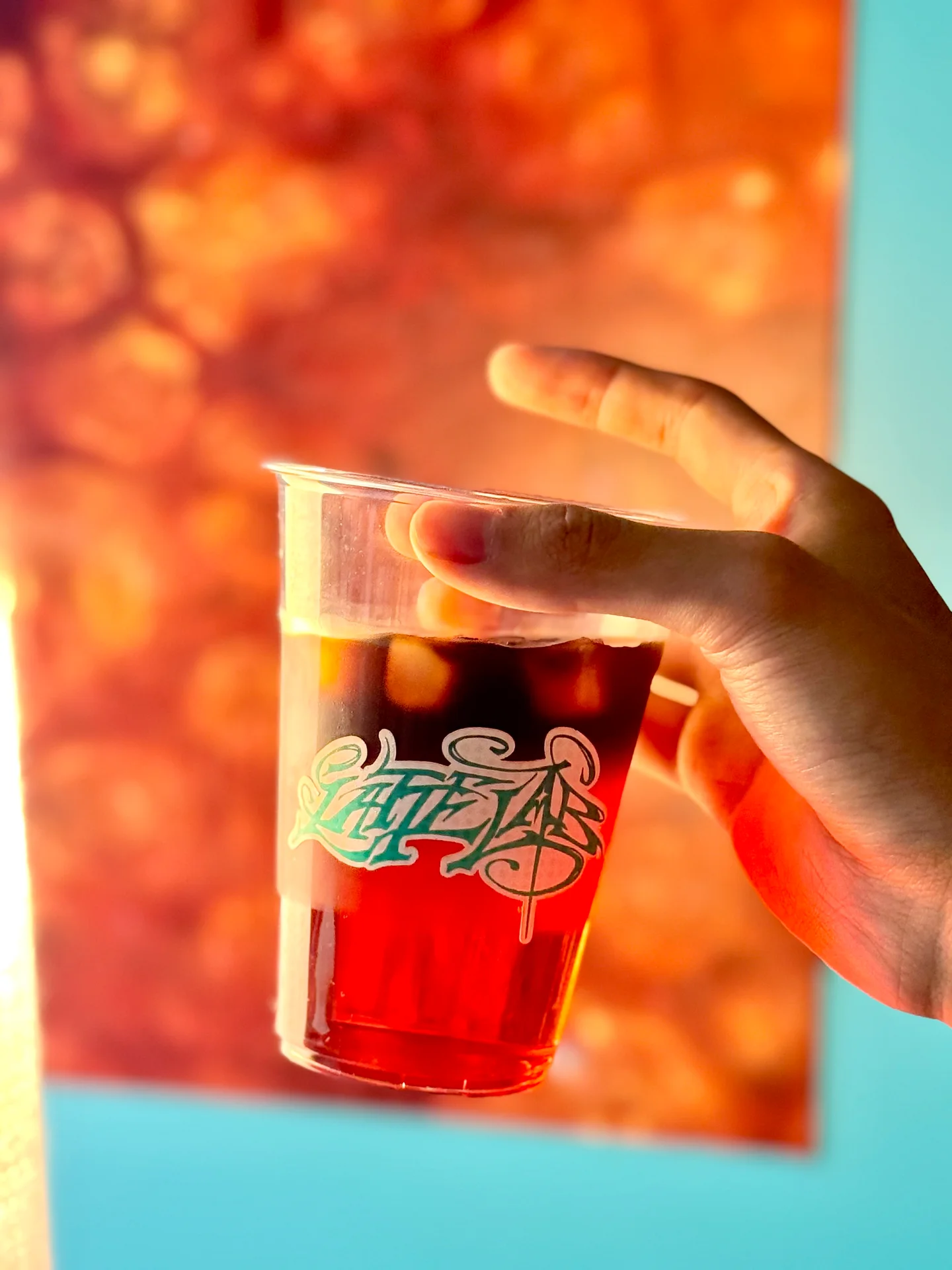 A hand holding passionfruit lemonade cold brew in a clear Latte Lab cup, backlit against orange and turquoise